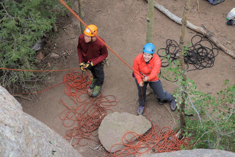 Two people are preparing for rock climbing. One person, wearing an orange helmet and a maroon sweater, is standing on the left. The other, wearing a blue helmet and an orange jacket, is on the right. Both are surrounded by coiled orange ropes, and they appear to be setting up their climbing gear near a rocky area with trees and foliage.